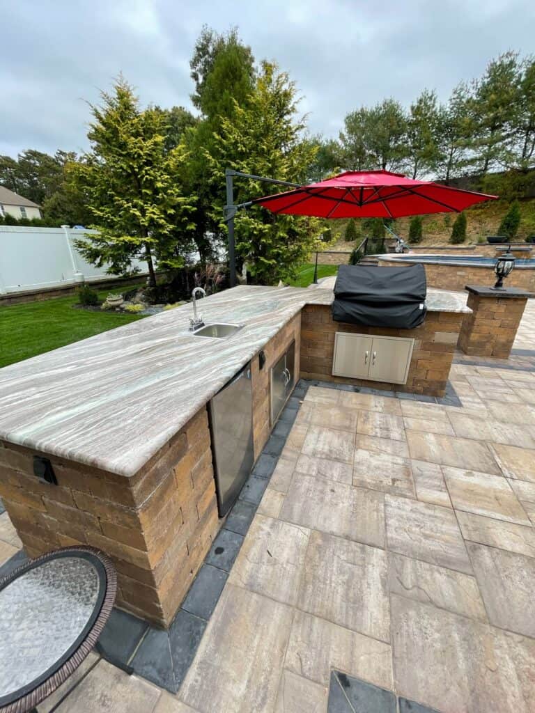 Outdoor kitchen featuring a stone countertop atop expert masonry craftsmanship. A built-in grill with a black cover and small sink accompany the setup. A red umbrella offers shade over the paved patio, framed by trees and a white fence typical of Suffolk County, NY.