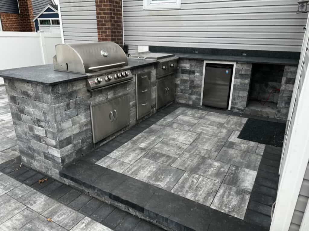 An outdoor kitchen featuring a stainless steel grill, cabinets, and mini fridge is built into expertly crafted masonry. Situated on a gray tiled patio in Suffolk County, NY, this setup includes a small mat adorning the space in front of the fridge.